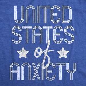May include: Blue t-shirt with the text "UNITED STATES of ANXIETY" in white, distressed lettering. Two white stars flank the word "of". The shirt has a classic, vintage style.