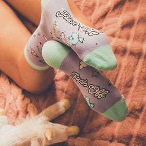May include: A pair of light purple crew socks with mint green heels and toes. The socks feature a repeating pattern of small green cacti and the text "Fuck Off" printed in a bold, dark, stylized script font across the top of the foot.
