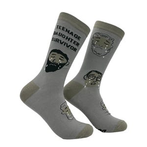 May include: A pair of gray socks with a humorous design. One sock features the text "TEENAGE DAUGHTER SURVIVOR" and a crying face with a beard. The other sock has two crying faces. The socks have olive green heels and toes.