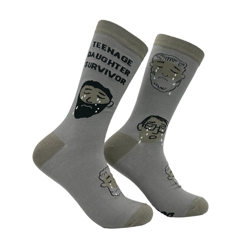 May include: A pair of gray socks with a humorous design. One sock features the text "TEENAGE DAUGHTER SURVIVOR" and a crying face with a beard. The other sock has two crying faces. The socks have olive green heels and toes.