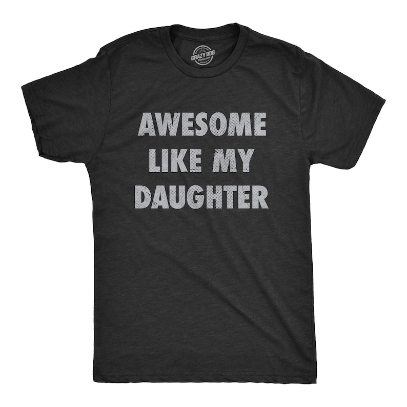 May include: A black t-shirt with the text "AWESOME LIKE MY DAUGHTER" in white, distressed lettering. The shirt has short sleeves and a crew neck. The Crazy Dog brand logo is visible at the top.