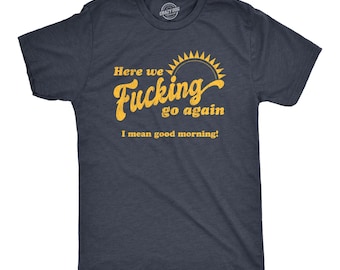 May include: A dark charcoal gray short-sleeve t-shirt featuring a yellow graphic print. The text reads "Here we Fucking go again" with a sun icon above, followed by the text "I mean good morning!" in a smaller, sans-serif yellow font.
