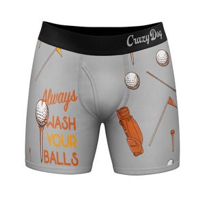 May include: Light gray boxer briefs with a black waistband featuring the text Crazy Dog. The fabric has an all-over print of golf balls, orange golf bags, and orange flags. The text Always WASH YOUR BALLS is printed in orange.