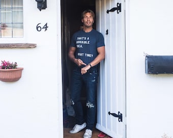 May include: A dark blue short-sleeved t-shirt with white text that reads, "THAT'S A HORRIBLE IDEA. WHAT TIME?" The shirt is paired with dark blue distressed denim jeans and white sneakers, creating a casual outfit for everyday wear.