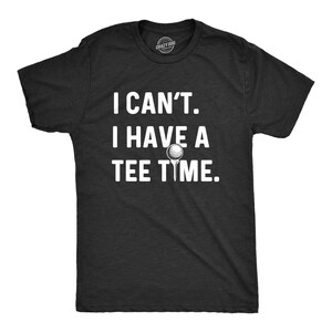 May include: A charcoal gray short-sleeved t-shirt featuring the white printed text I CAN'T. I HAVE A TEE TIME. The word TEE is illustrated with a small white golf ball resting on a golf tee, centered on the chest of the garment.
