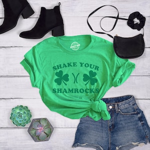 以下が含まれることがあります： 緑色のTシャツに「Shake Your Shamrocks」という文字と、緑色のクローバーのデザインが2つあります。Tシャツは前で結ばれており、デニムのショートパンツと合わせています。