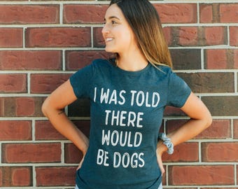 May include: A dark teal short-sleeved t-shirt featuring the white text "I WAS TOLD THERE WOULD BE DOGS" printed on the front. The shirt has a crew neckline and a fitted silhouette, paired with light blue denim jeans that have distressed details.