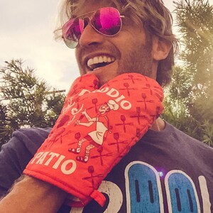 Op de afbeelding: Een persoon draagt een rode ovenwant met de tekst "Patio Daddy" en een witte en blauwe afbeelding. De ovenwant heeft een geruit patroon en een cartoonafbeelding van een persoon die een spatel vasthoudt.