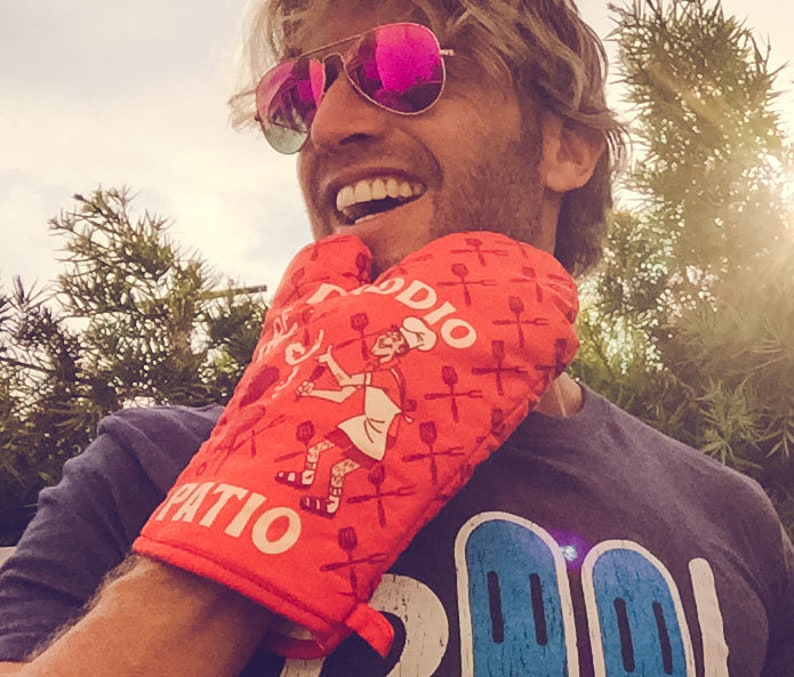 Op de afbeelding: Een persoon draagt een rode ovenwant met de tekst "Patio Daddy" en een witte en blauwe afbeelding. De ovenwant heeft een geruit patroon en een cartoonafbeelding van een persoon die een spatel vasthoudt.