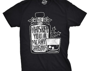 May include: A black short-sleeve t-shirt featuring a white graphic of a whiskey bottle and a glass with ice cubes. The text on the bottle reads WE WHISKEY YOU A MERRY CHRISTMAS, accompanied by illustrations of holly leaves and berries on the sides.