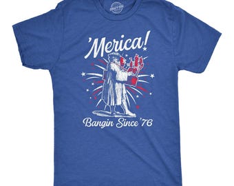 Może przedstawiać: Królewsko-niebieski t-shirt z krótkim rękawem z białym nadrukiem graficznym przedstawiającym postać historyczną trzymającą fajerwerki. Napis brzmi 'Merica! Bangin Since '76' białą czcionką, otoczony białymi i czerwonymi gwiazdami oraz fajerwerkami.