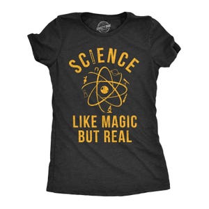 May include: A charcoal gray short-sleeve t-shirt featuring yellow distressed text that reads "SCIENCE LIKE MAGIC BUT REAL" with a central graphic of an atom surrounded by small scientific icons including a telescope, planet, and microscope.