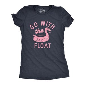 May include: A dark heather grey short-sleeve t-shirt featuring a light pink graphic of a flamingo pool float. The text "GO WITH the FLOAT" is printed in light pink above and below the flamingo illustration on the front of the garment.