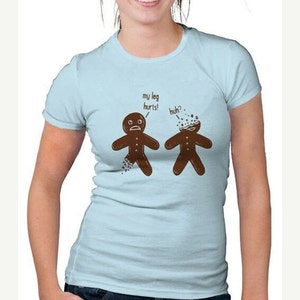 May include: Light blue t-shirt with a humorous graphic of two gingerbread men. One has a broken leg, the other a broken head. Text above reads "my leg hurts!" and "huh?". The shirt is worn by a person in blue jeans.