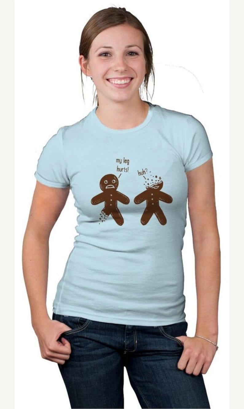 May include: Light blue t-shirt with a humorous graphic of two gingerbread men. One has a broken leg, the other a broken head. Text above reads "my leg hurts!" and "huh?". The shirt is worn by a person in blue jeans.