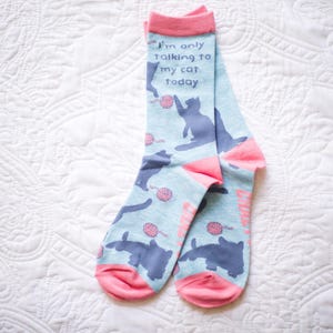 May include: A pair of light blue crew socks with a pink cuff, heel, and toe. The socks feature a repeating pattern of dark blue cat silhouettes and small pink yarn balls. The text "I'm only talking to my cat today" is printed in dark blue.