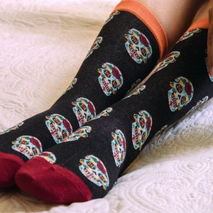 Könnte beinhalten: Ein Paar schwarze Socken mit einem bunten Totenkopf-Muster. Die Socken haben einen roten Bund und werden auf einem weißen Bettbezug getragen.