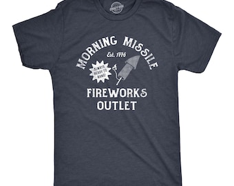 May include: A dark navy blue short-sleeve t-shirt featuring a white graphic print. The design includes the text "MORNING MISSILE", "Est. 1776", "ALWAYS READY TO BANG!", and "FIREWORKS OUTLET" alongside an illustration of a lit firework rocket.