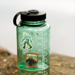 May include: A green water bottle with a black lid and two stickers. The stickers feature a cartoon Bigfoot in a forest setting. One sticker says "Hide & Seek Champion".