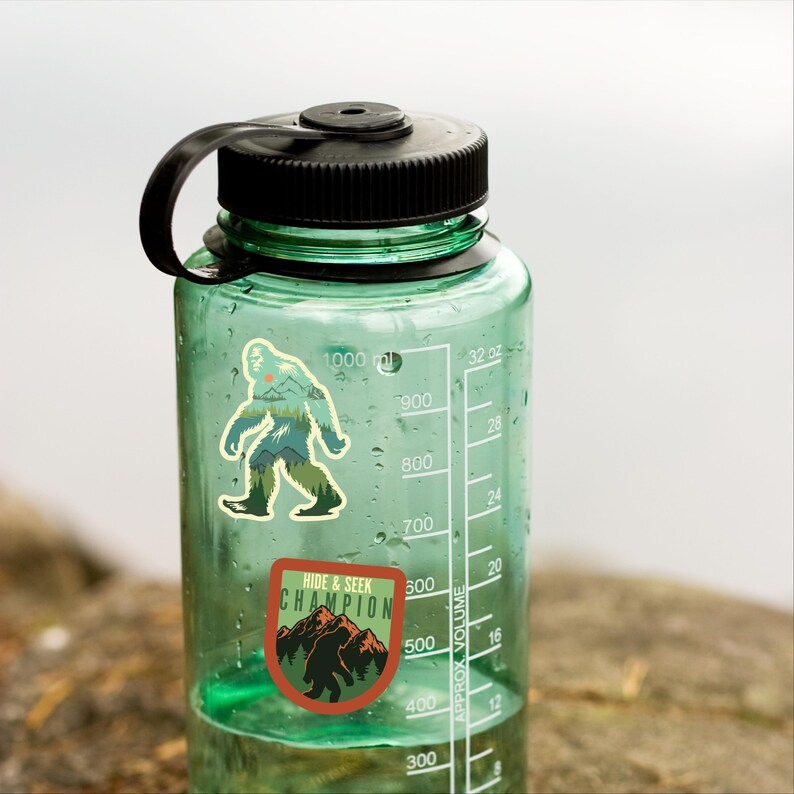 May include: A green water bottle with a black lid and two stickers. The stickers feature a cartoon Bigfoot in a forest setting. One sticker says "Hide & Seek Champion".