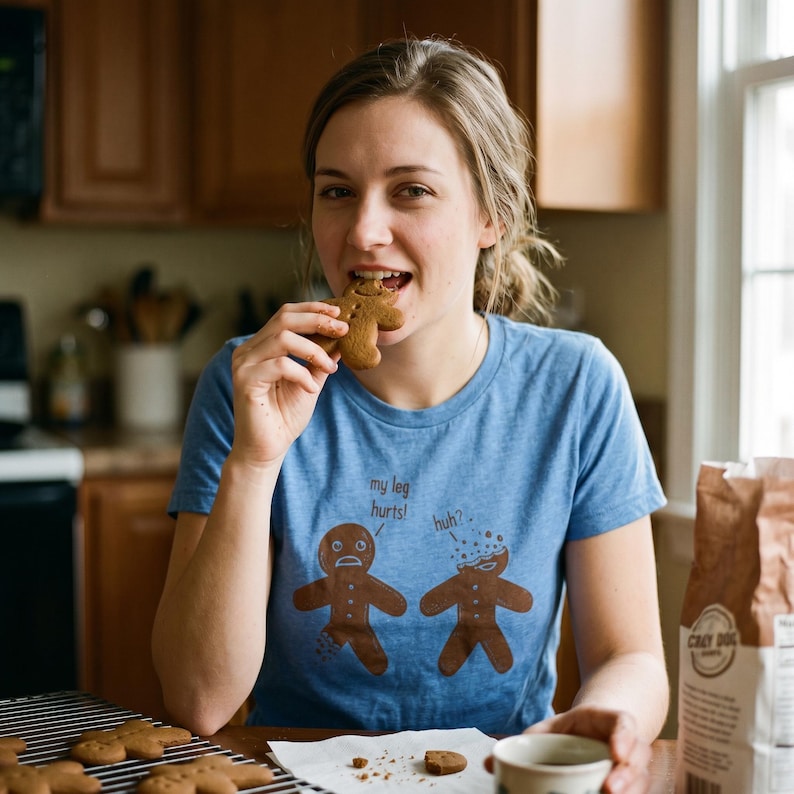 May include: A woman wearing a blue t-shirt with a gingerbread man graphic bites into a gingerbread cookie. The shirt reads "my leg hurts!" and "huh?". A bag of Crazy Dog flour is visible.