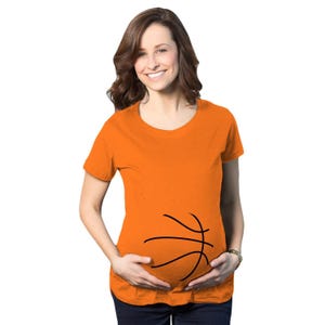 May include: A bright orange short-sleeved t-shirt featuring a black graphic print of basketball lines positioned across the midsection. The shirt has a round neckline and a simple, casual cut, designed to fit comfortably over a rounded belly area.