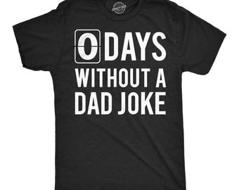 Op de afbeelding: Een zwart t-shirt met korte mouwen en witte tekst die 0 DAYS WITHOUT A DAD JOKE luidt, waarbij het cijfer 0 in een vierkant kader staat. Het shirt heeft een ronde hals en een standaard pasvorm met een duidelijke grafische print op de borst.