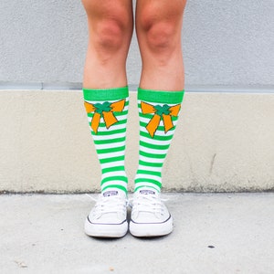 Puede incluir: Un par de zapatos de lona blancos con calcetines a rayas verdes y blancas con lazos naranjas con diseños de trébol.