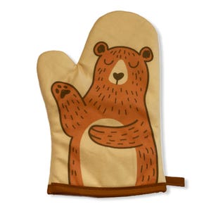 Può includere: Un guanto da forno beige con l'illustrazione di un orso marrone dal ventre color crema. L'orso ha gli occhi chiusi, il naso a forma di cuore e una zampa alzata. Il guanto è rifinito con un bordo marrone scuro e un piccolo occhiello laterale.