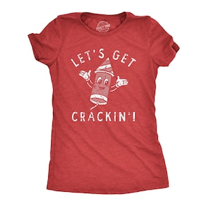 May include: A red short-sleeved t-shirt featuring a distressed white graphic of a cartoon firework character with arms outstretched. The text above the firework reads "LET'S GET" and the text below reads "CRACKIN'!" in a white, weathered font.