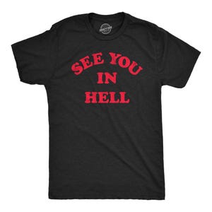 Op de afbeelding: Een zwart t-shirt met korte mouwen en de rode tekst SEE YOU IN HELL in een vet, verweerd lettertype op de borst gedrukt. Het kledingstuk heeft een ronde hals en een standaard pasvorm, met een klein merklogo zichtbaar in de kraag.