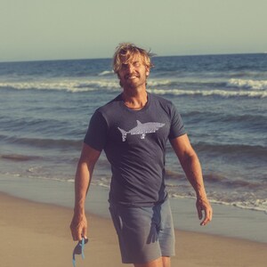 Puede incluir: Un hombre con una camiseta azul oscuro con un gr&aacute;fico de tibur&oacute;n blanco en la parte delantera. Est&aacute; de pie en una playa de arena con el oc&eacute;ano de fondo.