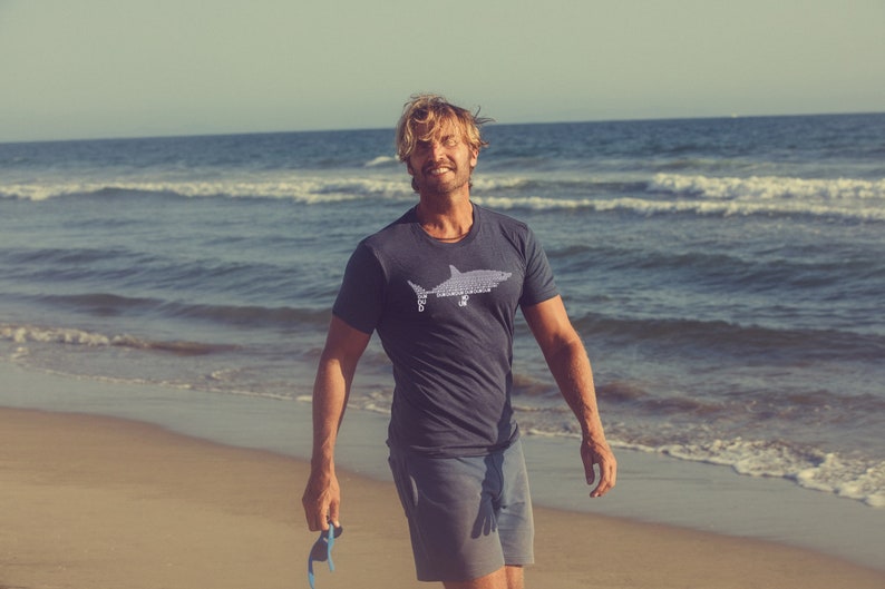 Puede incluir: Un hombre con una camiseta azul oscuro con un gr&aacute;fico de tibur&oacute;n blanco en la parte delantera. Est&aacute; de pie en una playa de arena con el oc&eacute;ano de fondo.
