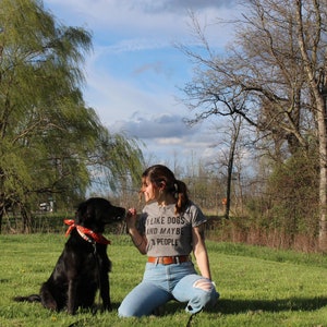 May include: A black dog with an orange bandana sits on green grass next to a person wearing a gray t-shirt with the text "I LIKE DOGS AND MAYBE 3 PEOPLE". The dog and person are outside on a sunny day.