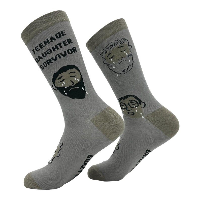 May include: A pair of gray socks with the text "TEENAGE DAUGHTER SURVIVOR" and illustrations of crying faces. The socks have a gray heel and toe, and the word "CRAZY" is printed on the sole.