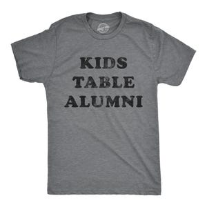 Könnte beinhalten: Graues T-Shirt mit dem Text "KIDS TABLE ALUMNI" in schwarzer, verwaschener Schrift. Das Shirt hat einen Rundhalsausschnitt und kurze Ärmel. Das Crazy Dog-Logo ist am Ausschnitt zu sehen.