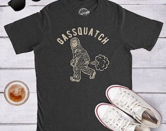 Può includere: Una t-shirt a maniche corte grigio antracite con la grafica di una creatura pelosa che cammina ed emette una nuvola di gas. Sopra la creatura, il testo recita GASSQUATCH in un carattere chiaro e dall'aspetto usurato. Girocollo.