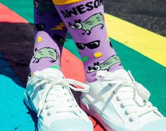 May include: Purple crew socks with a yellow cuff featuring a repeating pattern of green opossums wearing black sunglasses, accompanied by the text AWESOME in black letters, paired with white lace-up canvas sneakers with light blue rubber toes.