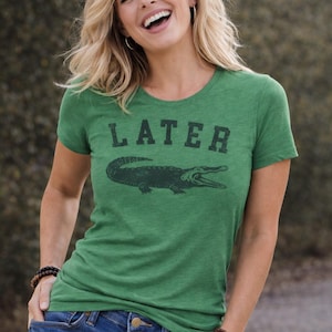 May include: Green t-shirt with the word "LATER" above a dark green alligator graphic. The shirt is a crew neck style and is paired with blue denim jeans. The model is smiling.
