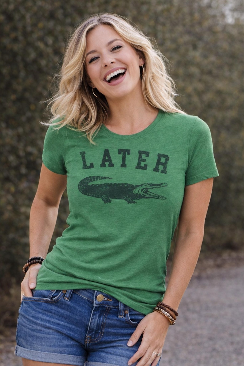 May include: Green t-shirt with the word "LATER" above a dark green alligator graphic. The shirt is a crew neck style and is paired with blue denim jeans. The model is smiling.