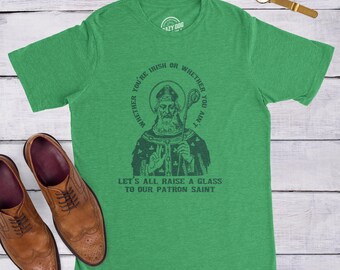 Puede incluir: Una camiseta de manga corta de color verde brillante con un gráfico en verde oscuro de un santo sosteniendo un báculo. El texto dice: WHETHER YOU'RE IRISH OR WHETHER YOU AIN'T LET'S ALL RAISE A GLASS TO OUR PATRON SAINT. La camiseta se muestra con zapatos de cuero marrón.