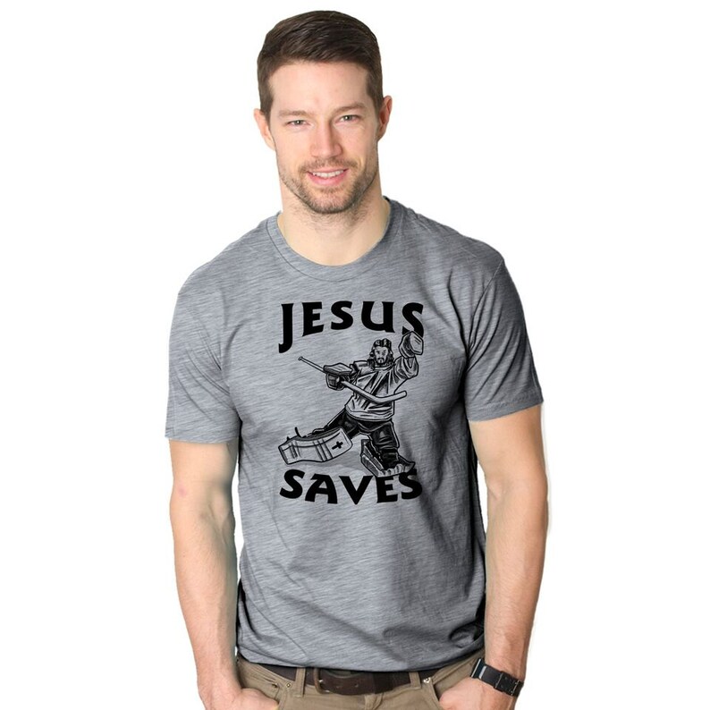 Puede incluir: Una camiseta gris jaspeada con un gr&aacute;fico negro de Jes&uacute;s jugando al hockey. El texto "JESUS SAVES" est&aacute; impreso debajo del gr&aacute;fico.