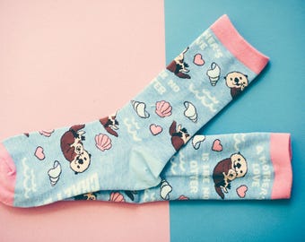 May include: Light blue crew socks with pink cuffs, heels, and toes. The pattern features brown otters, pink hearts, white seashells, and wavy lines. The text reads A MOTHER'S LOVE IS LIKE NO OTTER, printed in white capital letters across the fabric.