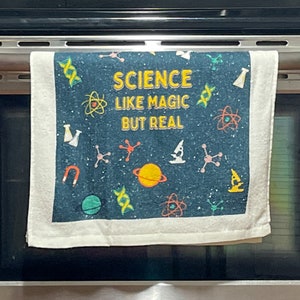 May include: A white kitchen towel with a blue background and colourful science-themed illustrations. The text "SCIENCE LIKE MAGIC BUT REAL" is printed in yellow on the towel.