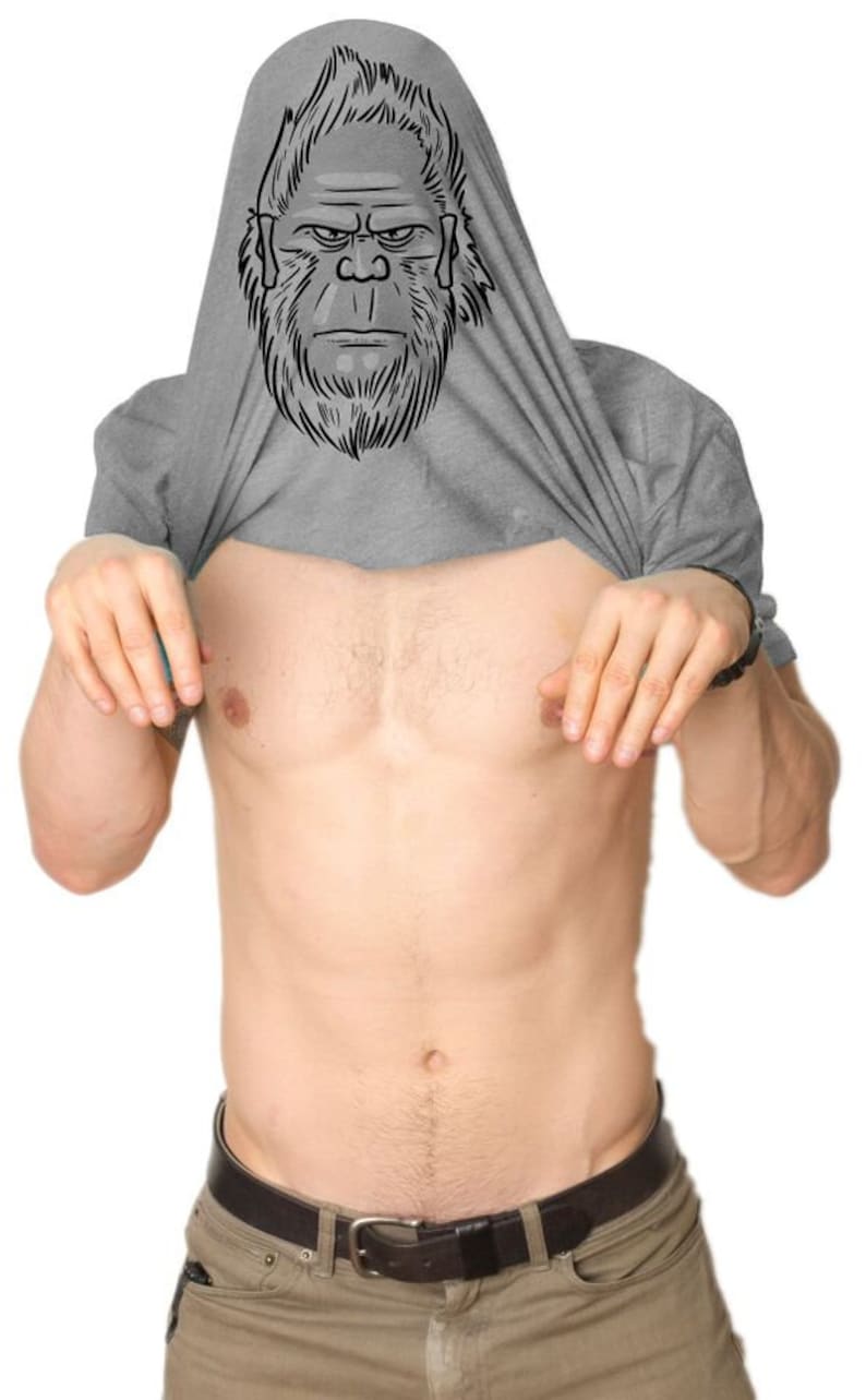 May include: A gray t-shirt with a black and white illustration of a Bigfoot's face on the front. The shirt is pulled up over the person's head to reveal the illustration.