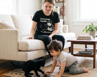 May include: A black short-sleeved t-shirt featuring a white graphic illustration of a child and a cat. The text on the shirt reads "Everyone Sucks" above the illustration and "EXCEPT FOR THIS FUCKING AWESOME CAT" below the illustration in white lettering.