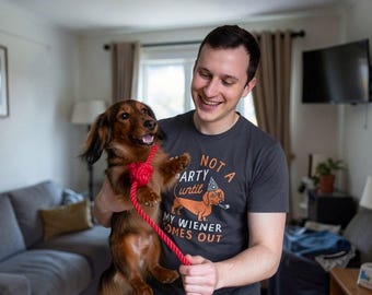 May include: A charcoal gray t-shirt featuring a graphic of a dachshund wearing a party hat with the text NOT A PARTY until MY WIENER COMES OUT. The shirt is paired with a long-haired brown dachshund wearing a bright red rope leash.