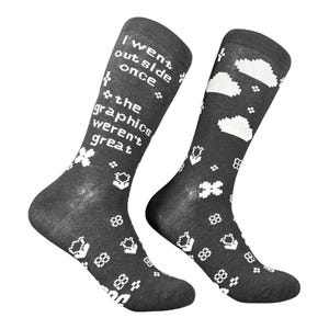 Puede incluir: Un par de calcetines gris oscuro con gráficos pixelados blancos. Un calcetín muestra el texto "I went outside once the graphics weren't great" en una fuente retro, mientras que el otro presenta nubes pixeladas blancas y varias formas geométricas pequeñas.