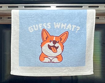 Puede incluir: Un paño de cocina azul claro con un borde blanco y un gráfico central de un perro Corgi marrón y blanco con la lengua fuera. Sobre el perro, el texto GUESS WHAT? está impreso en letras mayúsculas blancas, negritas y sin remates.