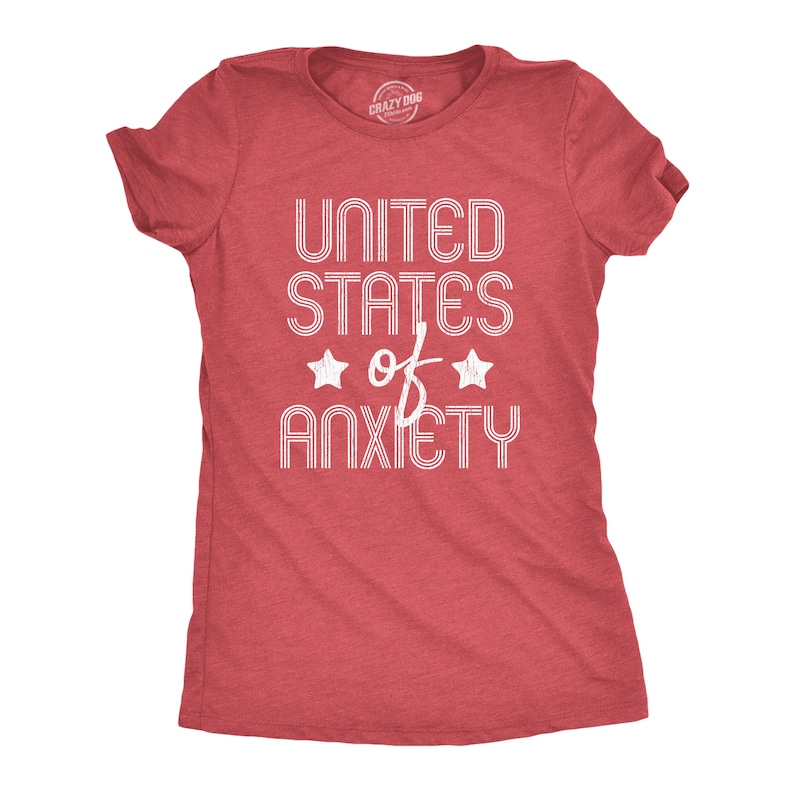 May include: A red t-shirt with the text "UNITED STATES of ANXIETY" in white, stylized font. Two white stars flank the word "of". The shirt is a crew neck style.